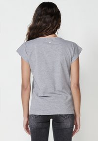 Grå T-shirt med korta ärmar, något rundad halsringning och en liten logotyp på övre ryggen, gjord av mjukt, lätt tyg.