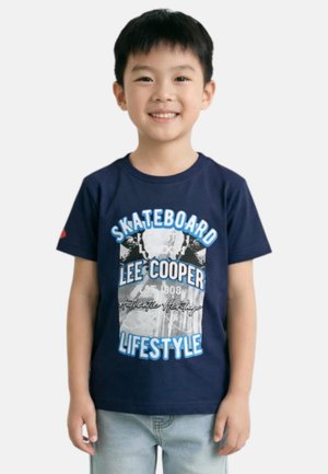 Garçon souriant portant un t-shirt graphique bleu marine "Skateboard Lee Cooper Lifestyle" et un jean bleu clair, debout devant un fond blanc.