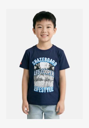 Garçon souriant portant un t-shirt graphique bleu marine "Skateboard Lee Cooper Lifestyle" et un jean bleu clair, debout devant un fond blanc.