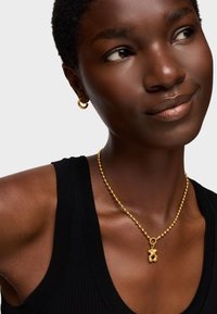 Collana di perline d'oro con chiusura e un ciondolo decorativo, abbinata a orecchini a cerchio d'oro. Il modello indossa un top nero senza maniche.