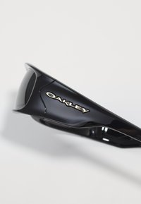 Juodos Oakley saulės akinių šakelės su baltu logotipu, artimas planas sklandžiai, blizgiai plastikinei rėmeliui prieš šviesų foną.