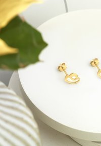 Goldene Ohrringe mit einer offenen Herzform und Steckverschluss, präsentiert auf einer weißen runden Plattform mit umliegendem grünem Laub.