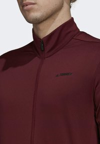 Chaqueta deportiva burdeos con cuello alto y cierre de cremallera; presenta el logo negro "TERREX" en el pecho; textura de tejido suave y elástico.