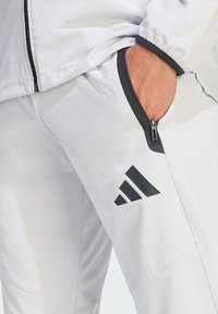 Białe spodnie sportowe z czarnymi akcentami, wyposażone w kieszeń na zamek i logo Adidas. Gładka faktura, zwężany krój i lekki materiał.