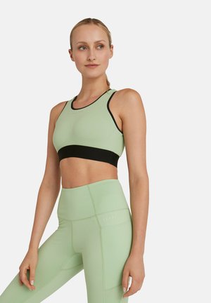 Frau trägt ein hellgrünes Sport-BH mit schwarzer Einfassung und passende hoch taillierte Leggings, steht mit einer Hand auf dem Oberschenkel.