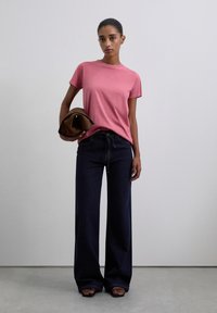 T-shirt rose à manches courtes associé à un jean en denim bleu foncé taille haute et large. La personne tient une pochette marron et porte des sandales à lanières.