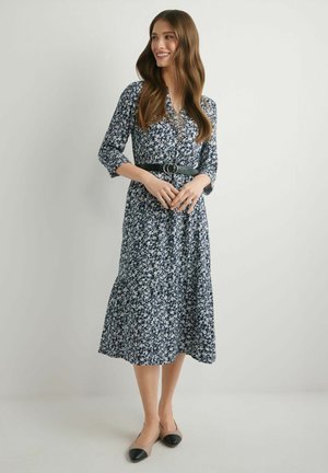 Kobieta uśmiechająca się, ubrana w granatową sukienkę midi w kwiaty z rękawami 3/4, czarny pasek z podwójną klamrą i beżowe dwukolorowe baleriny, stojąca na jednolitym tle.