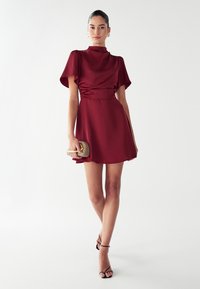 Burgundi spalvos suknelė su prigludusia juosmuo, pūstomis trumpomis rankovėmis ir lygiu audiniu. Sukurta su mažą auksiniu akcentu panku ir juodais sandalais su juostelėmis.