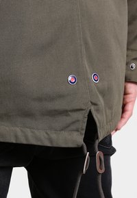 Veste vert olive en tissu texturé, dotée de deux boutons-pression en métal avec des accents bleus et rouges à l'ourlet et d'une fermeture à cordon.