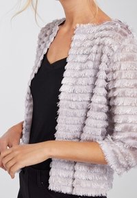 Chaqueta de textura púrpura clara con diseño de flecos en capas, longitud corta y delantero abierto, llevada sobre una blusa negra.