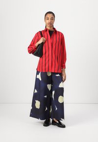Camicia a righe rosse con bottoni abbinata a pantaloni larghi blu navy con un motivo astratto crema. Gli accessori includono una borsa nera e dei mocassini.