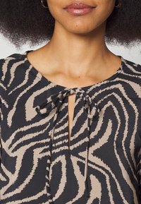 Blusa preta e bege com um padrão zigzag texturizado, decote redondo e um detalhe laçado à frente. Tecido suave com um corte descontraído.