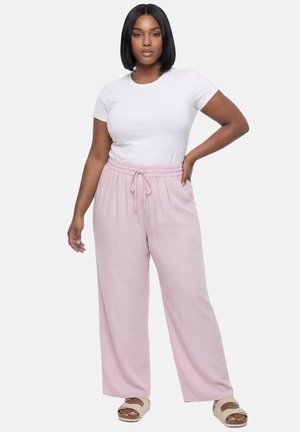 Kobieta stoi z ręką na biodrze, ubrana w biały krótki rękaw, jasnoróżowe spodnie na sznurku i beżowe klapki na białym tle.