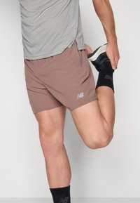 Sportshorts aus muted braunem Stoff, mit einem elastischen Bund, Seitentaschen und kleinem silbernen Branding. Kombiniert mit einem grauen Oberteil und schwarzen Socken.