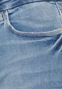 Vaqueros de mezclilla azul con un acabado desgastado, presentan un solo bolsillo y detalles de costura visibles. La tela parece suave con un ligero brillo.