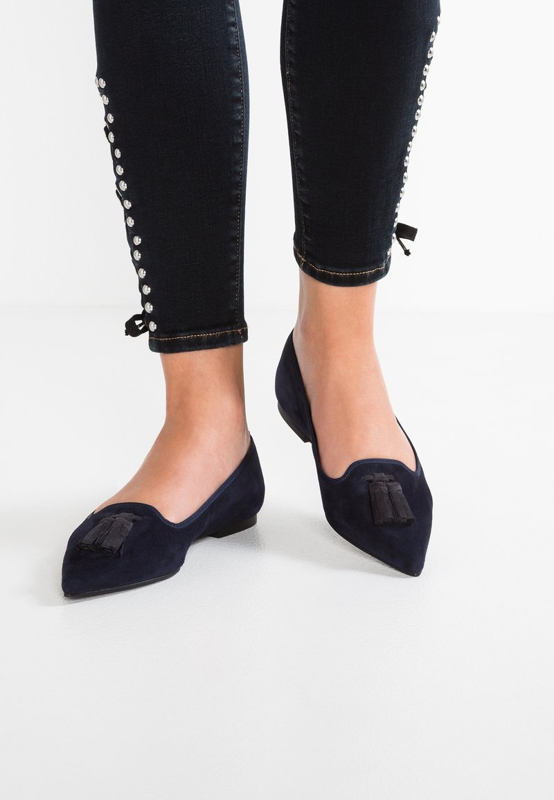 Pieds en mocassins noirs à bouts pointus avec des glands, portant un pantalon court noir orné de clous argentés sur les côtés.