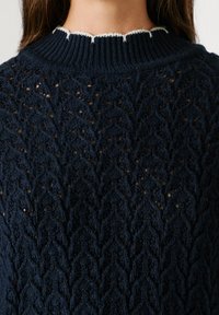 Tumesinine kootud sviiter, millel on tekstuuriga zigzag-muster, ribikraega, millel on kontrastne valge äär, ning lõtv ja hingav kangas.