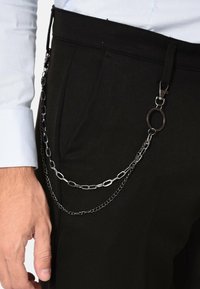 I pantaloni neri presentano una catena in metallo argento attaccata a un passante della cintura, con una combinazione di anelli rotondi e ovali, e una clip per fissare oggetti.