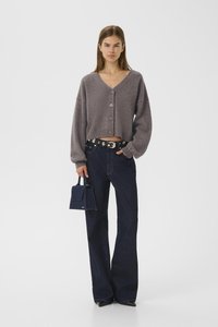 Woman wearing a gray buttoned cardigan, dark wide-leg jeans with a black studded belt, black pointed shoes, and holding a small navy handbag.