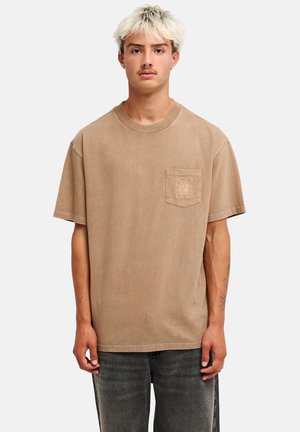 Jonge man met kort gebleekt haar, draagt een loszittend beige T-shirt met een klein zakje en zwarte uitgevaagde jeans, staand tegen een effen achtergrond.