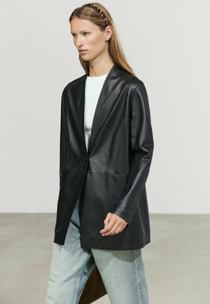 Kvinde iført en sort læderblazer over en hvid top og lyseblå jeans med leopardprint hårbånd, stående mod en ensfarvet baggrund.