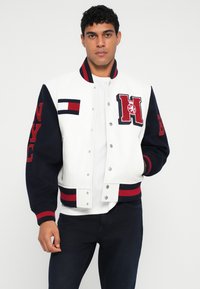 Giacca varsity bianca con maniche blu navy, caratterizzata da scritte rosse "NYC" e "H". Polsini e vita a coste con strisce orizzontali. Chiusure a scatto metalliche.
