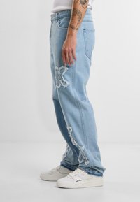 Lyseblå denimjeans med slitte lapper, med store hvite bokstaver, løs passform, og kombinert med hvite joggesko.