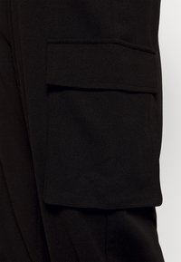 Tissu de pantalon noir à la texture lisse, doté d'une grande poche cargo plate sur le côté. Le design est minimaliste et utilitaire.
