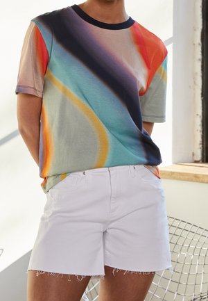 Osoba ubrana w krótkim rękawem t-shirt z wielokolorowymi przekątnymi paskami i białe szorty z postrzępionym brzegiem, stojąca w pomieszczeniu przy oknie.