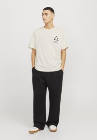Camiseta crema de manga corta con un logo en forma de triángulo negro y texto en el lado izquierdo del pecho, combinada con pantalones negros holgados y zapatillas blancas.