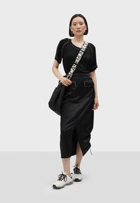 Černý outfit zahrnující top s krátkým rukávem s hroty a hladkou sukni s vysokým rozparkem. V kombinaci s bílými teniskami a černou taškou.