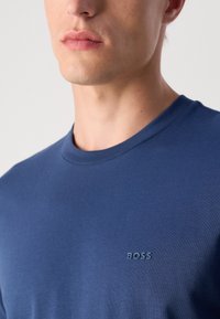 Acercamiento a un hombre que lleva una camiseta de cuello redondo azul marino con un pequeño logo "BOSS" en la parte izquierda del pecho.