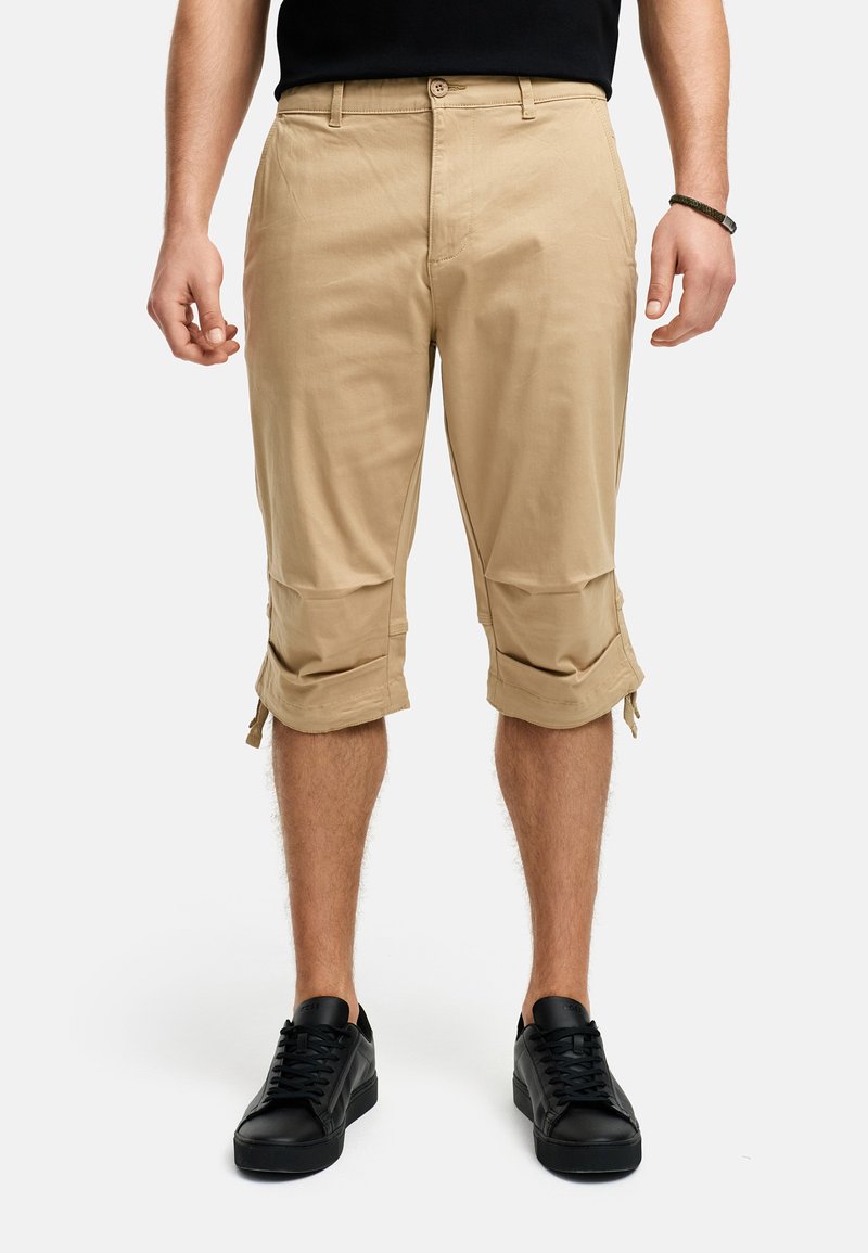 Shorts cargo beiges avec ourlets à revers, deux poches latérales et fermeture à bouton, associés à des baskets noires sur un fond neutre.
