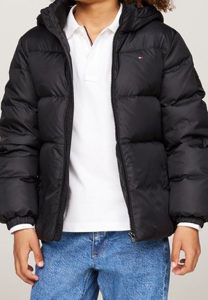 Enfant portant une doudoune noire par-dessus un polo blanc et un jean bleu, debout avec les mains détendues sur les côtés.
