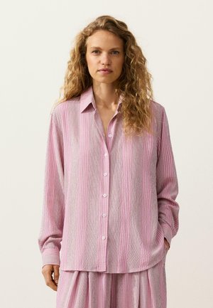 Kvinde med krøllet hår iført løs skjorte med pink og hvide striber og matchende bukser med vid ben mod en ensfarvet baggrund.