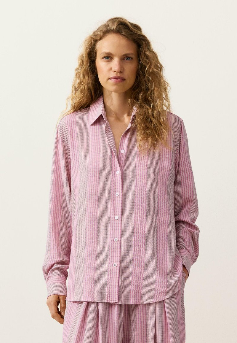 Femme aux cheveux bouclés portant une chemise ample à rayures roses et blanches avec des boutons, ainsi qu'un pantalon large assorti, sur un fond uni.