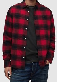 Camicia a quadri rossa e nera in tessuto di flanella con chiusura frontale con bottoni, bottoni bianchi e logo sul petto. Indossata sopra una t-shirt nera.