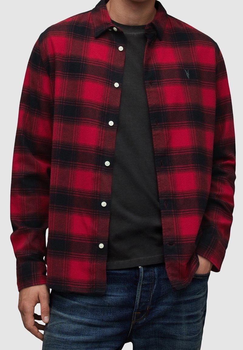 Camicia a quadri rossa e nera in tessuto di flanella con chiusura frontale con bottoni, bottoni bianchi e logo sul petto. Indossata sopra una t-shirt nera.