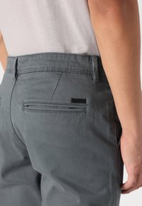 Chinos grises con una textura suave que cuentan con un bolsillo trasero con una etiqueta negra. La cinturilla tiene trabillas para el cinturón y un acabado limpio.
