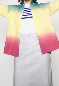 Nappikauluksinen paita liukuvärjäyksellä: keltainen vihreän kautta vaaleanpunaiseen, yhdistettynä valkoiseen T-paitaan, jossa on sinisiä raitoja, sekä vaaleanharmaaseen hameeseen.