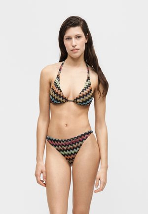 Fiatal nő hosszú barna hajjal előre néz, és egy többszínű cikkcakk mintás, nyakpántos bikiniben áll, egyszerű háttér előtt.