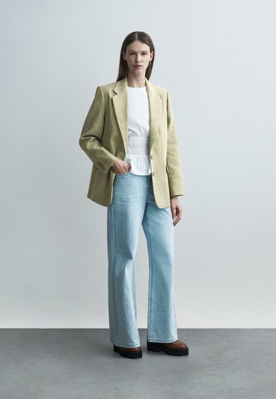 Femme portant un blazer beige à fines rayures, un chemisier blanc à taille smockée, un jean large bleu clair orné de décorations, et des chaussures compensées marron.