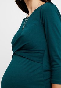 Miętowa bluzka z elastycznego materiału z rękawami o długości trzech czwarte, z twistowanym wzorem w talii, idealna na ubrania ciążowe.