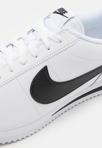 White sneaker with textured black Nike logo, lace-up design, smooth synthetic upper, and white rubber sole with zigzag pattern.