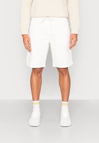 Shorts blancs en coton avec une ceinture élastique, fermeture à cordon, et poches latérales, accompagnés de baskets blanches et de chaussettes rayées.