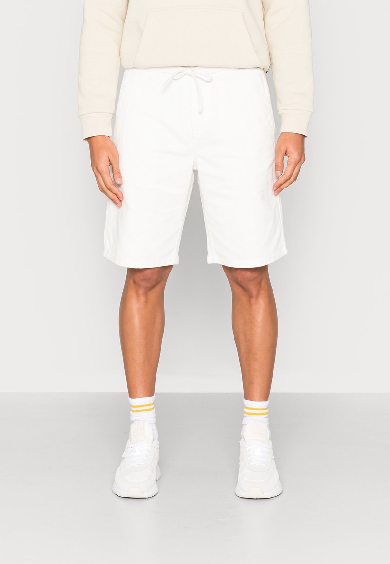 Shorts blancs en coton avec une ceinture élastique, fermeture à cordon, et poches latérales, accompagnés de baskets blanches et de chaussettes rayées.