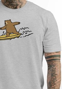 Graues T-Shirt mit kurzen Ärmeln, das einen Cartoon-Bären zeigt, der auf einem gelben Brett surft, mit schwarzen Umrisswellen darunter.