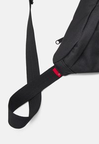 Borsa a tracolla nera in tessuto con superficie testurizzata, dotata di una chi strap regolabile ampia e un'etichetta d'accento rossa vicino al punto di connessione.