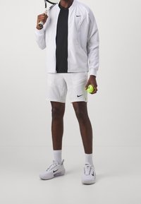 Man i vit sportklädsel som håller en tennisracket på axeln och en gul tennisboll, står framför en enfärgad bakgrund.