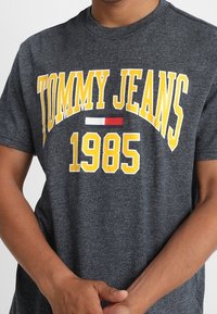 Camiseta de algodón gris con mangas cortas que presenta un audaz texto amarillo "TOMMY JEANS" y "1985" en una franja blanca con acentos en rojo y azul.